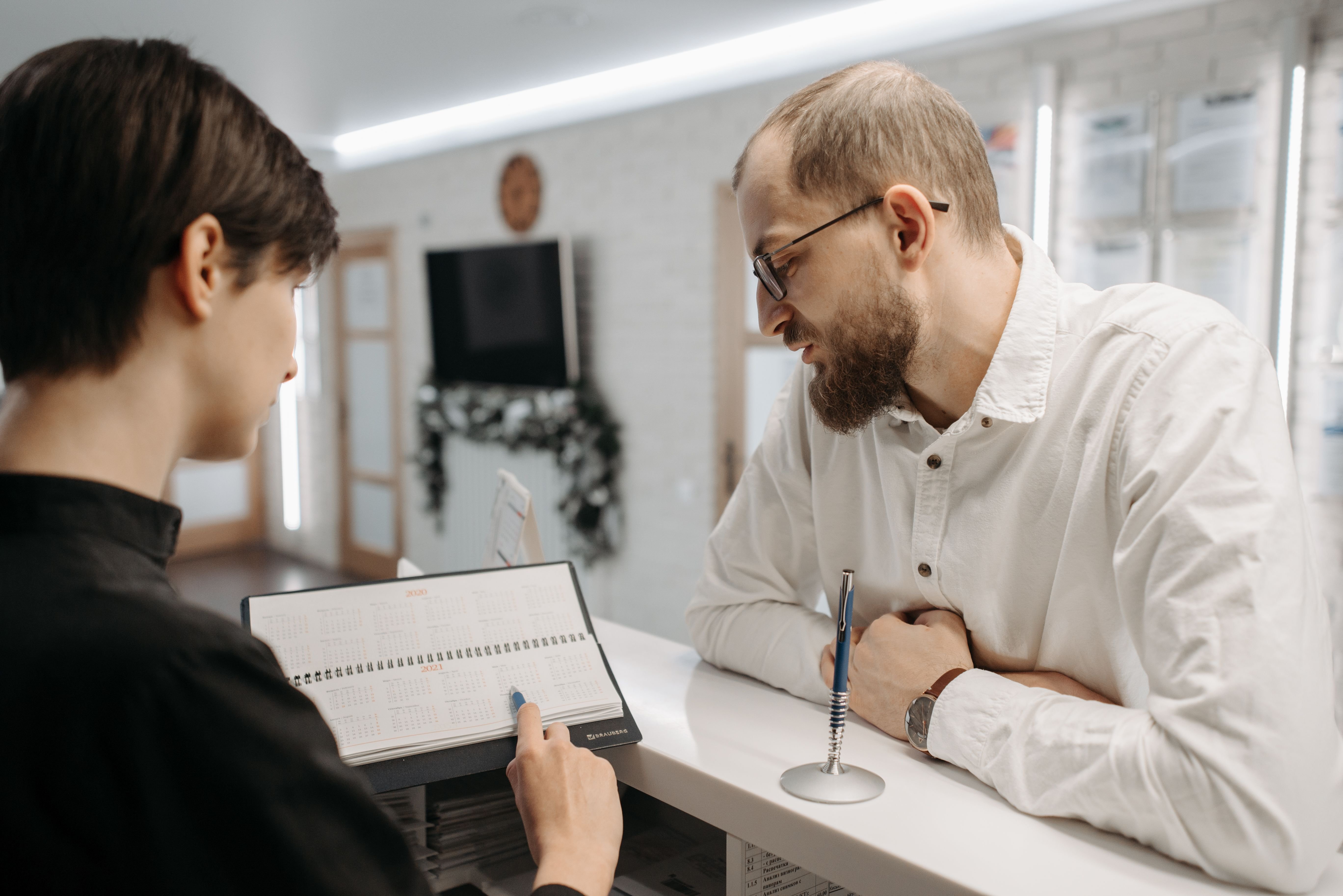 A receptionist and a patient at a clinic front desk, looking at an appointment diary together.