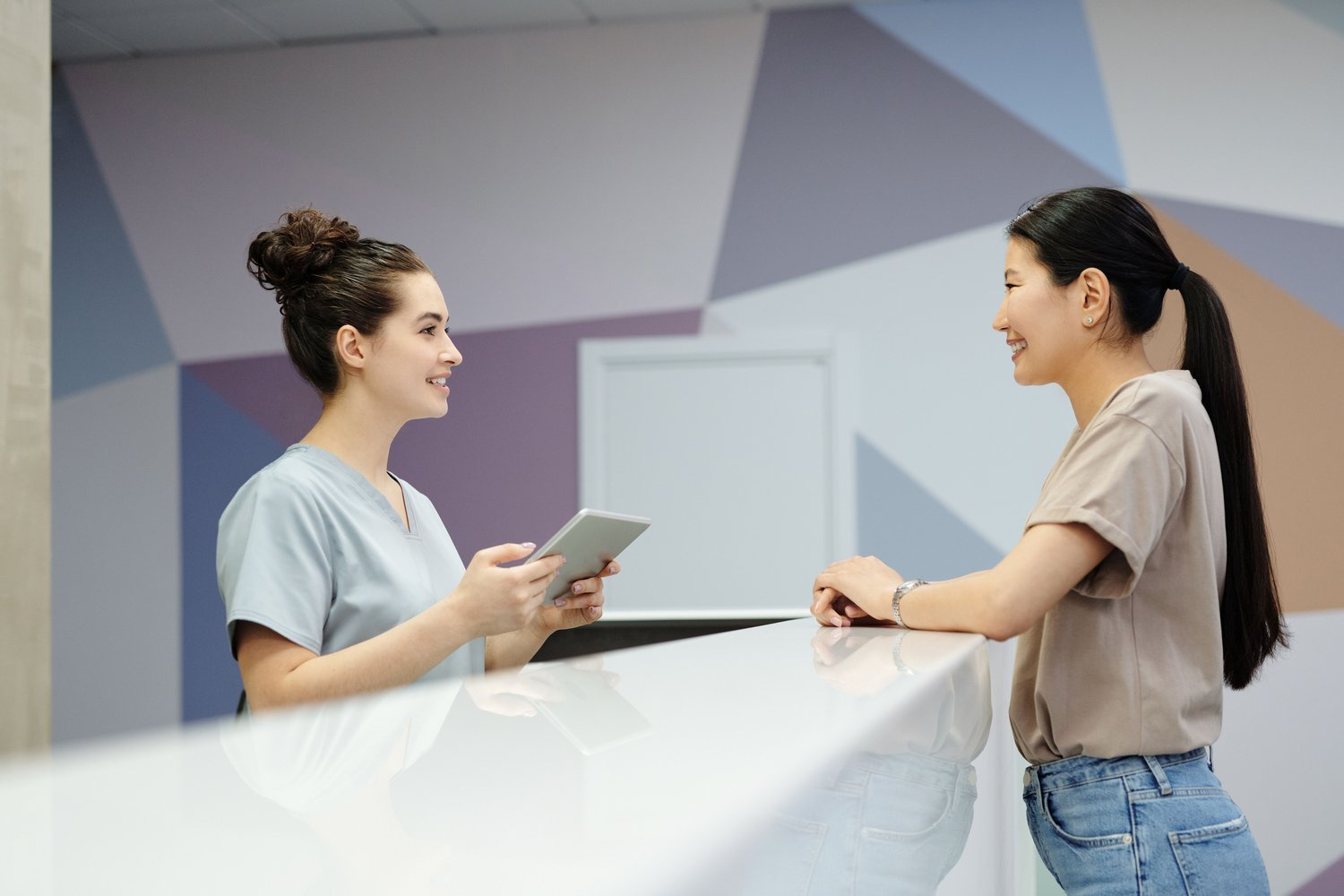 A practitioner with a tablet talking to a patient across a clinic reception desk