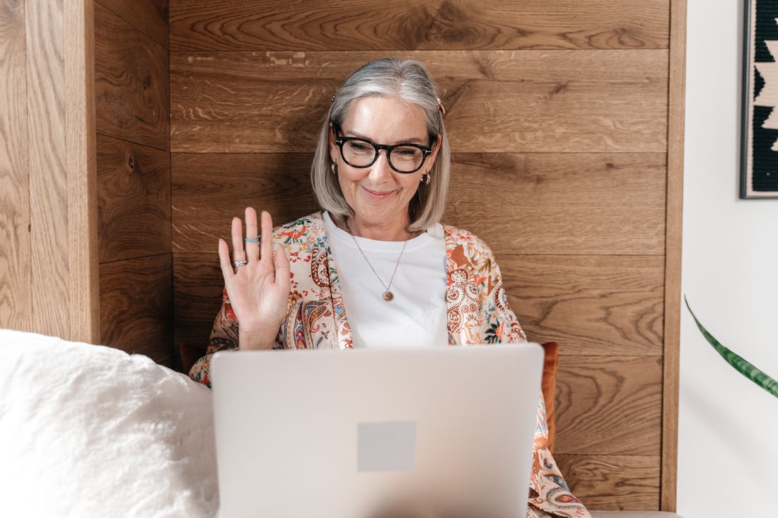 A woman sitting at home, waving at the laptop in front of her as a telehealth consult starts.