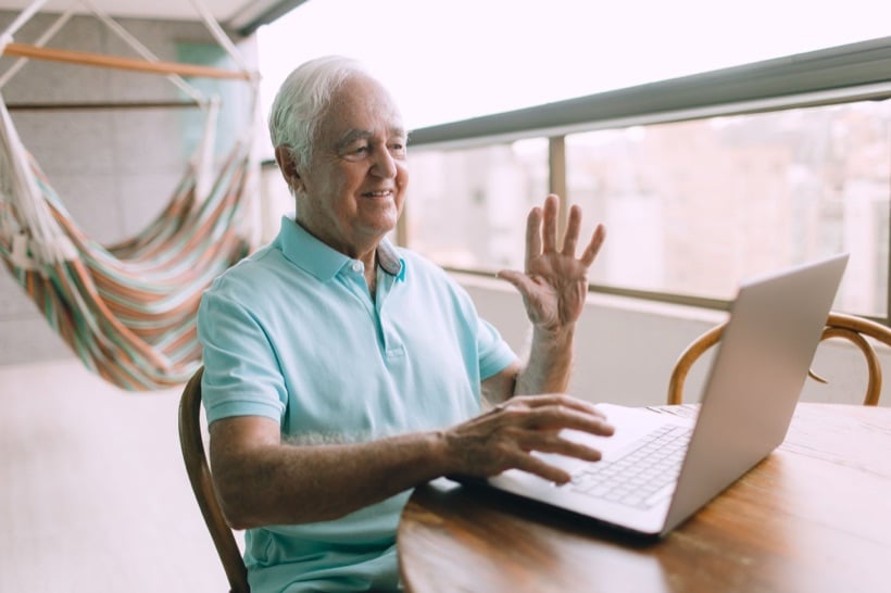 An older patient smiling and waving hello as he joins a telehealth appointment on his laptop