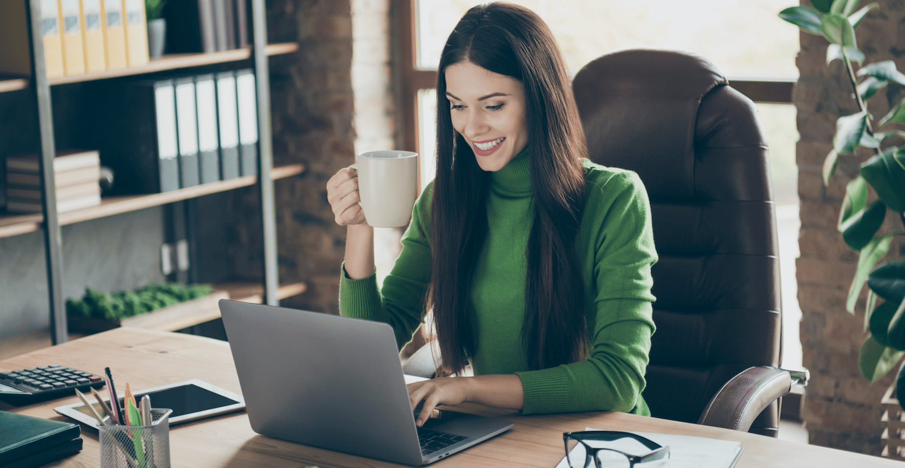 Woman in home office sits by computer whilst holding a cup of tea